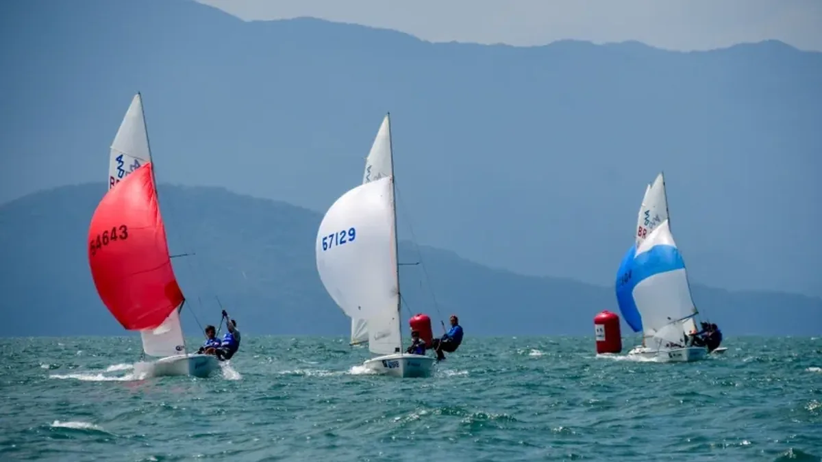 Três veleiros, um de vela vermelha, um de vela branca e um de vela azul e branca, navegando em frente às montanhas