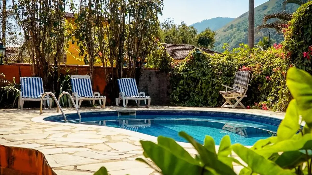 Piscina de pousada cercada de espreguiçadeiras e jardim tropical, com montanhas ao fundo